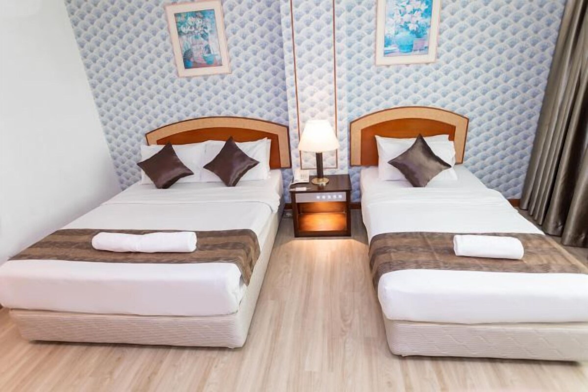 A hotel room with two beds, each with a wooden headboard, white bedding, and a folded towel on the bed. There is a nightstand with a lamp between the beds. The walls have a textured pattern, and there are two framed pictures above the beds.