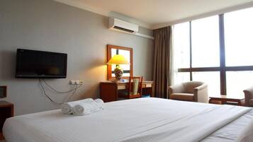 A hotel room with a large bed, a desk with a lamp, a television, and a large window with curtains.