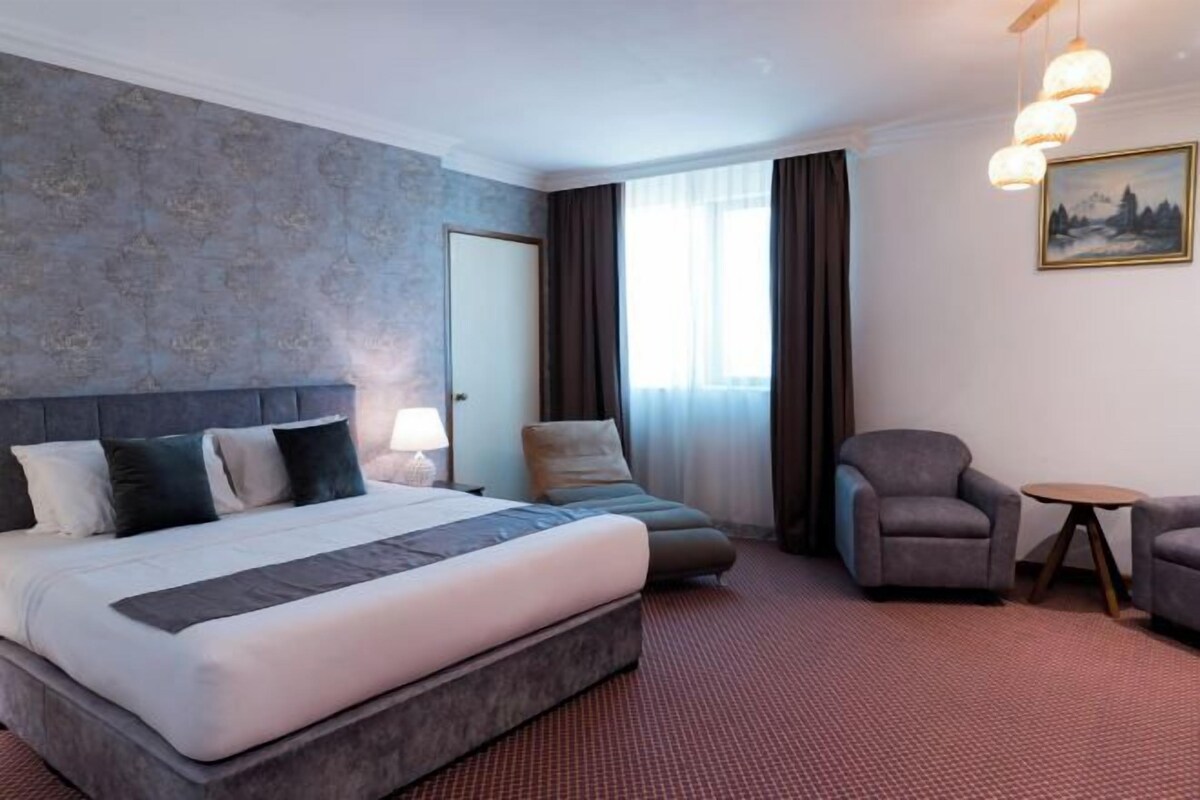 A modern hotel room with a large bed, a sofa, two armchairs, a small table, and a painting on the wall.