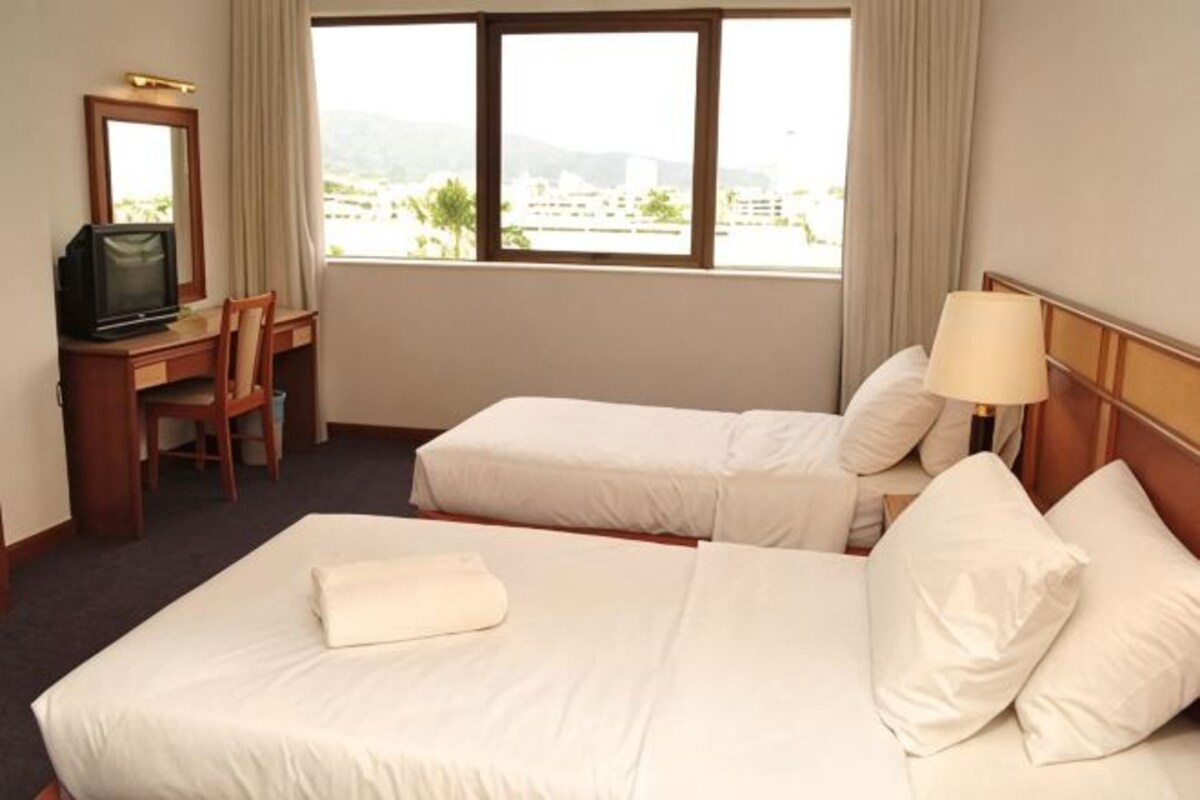A hotel room with two beds, a desk with a television, and a window with a view of buildings and hills.