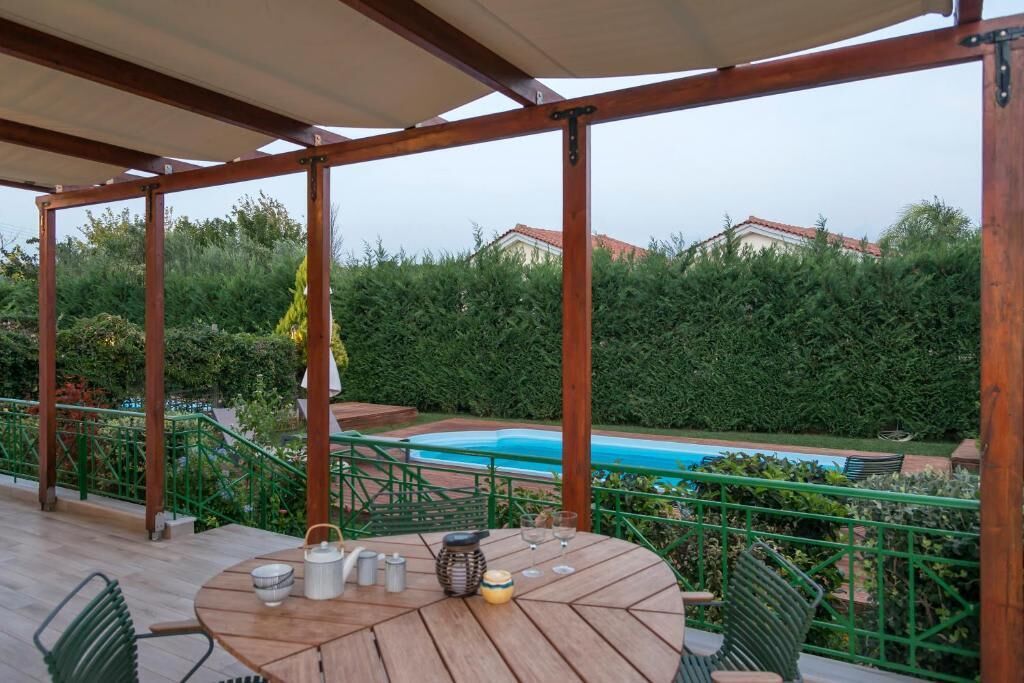 A wooden deck with a round table and chairs, a pool in the background, and a green hedge surrounding the area.