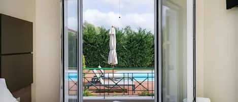 Habitación de hotel con cama, mesita de noche, televisor y balcón con vistas a una piscina y zonas verdes.