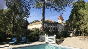 A swimming pool with lounge chairs, surrounded by trees and a historic building with a red-tiled roof.