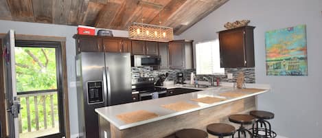 A modern kitchen with a wooden ceiling, stainless steel appliances, a central island with stools, and a view through a glass door.