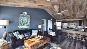 Uma sala de estar moderna com paredes azul-escuras, teto de madeira e área de cozinha visível por uma porta aberta.
