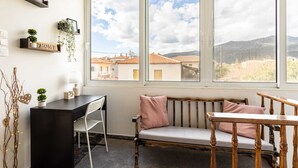 A room with a desk, a bench, and a large window offering a view of buildings and mountains. - Sunset Serenity - Kardamili Seaside Getaway (Kardamyli)