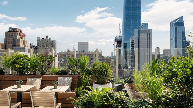 A rooftop terrace with wooden furniture and potted plants, overlooking a city skyline with modern buildings.