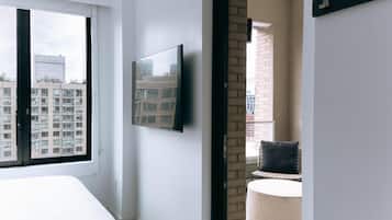 A modern hotel room with a large window, a flat-screen TV mounted on the wall, a round seating area, and a view of the cityscape.