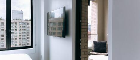 A modern hotel room with a large window, a flat-screen TV mounted on the wall, a round seating area, and a view of the cityscape.