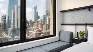 A modern hotel room with a large window offering a city view, a comfortable seating area, and a bedside table with a plant.