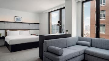 Habitación de hotel moderna con cama, sofá, escritorio y ventanas amplias con vista a la ciudad.