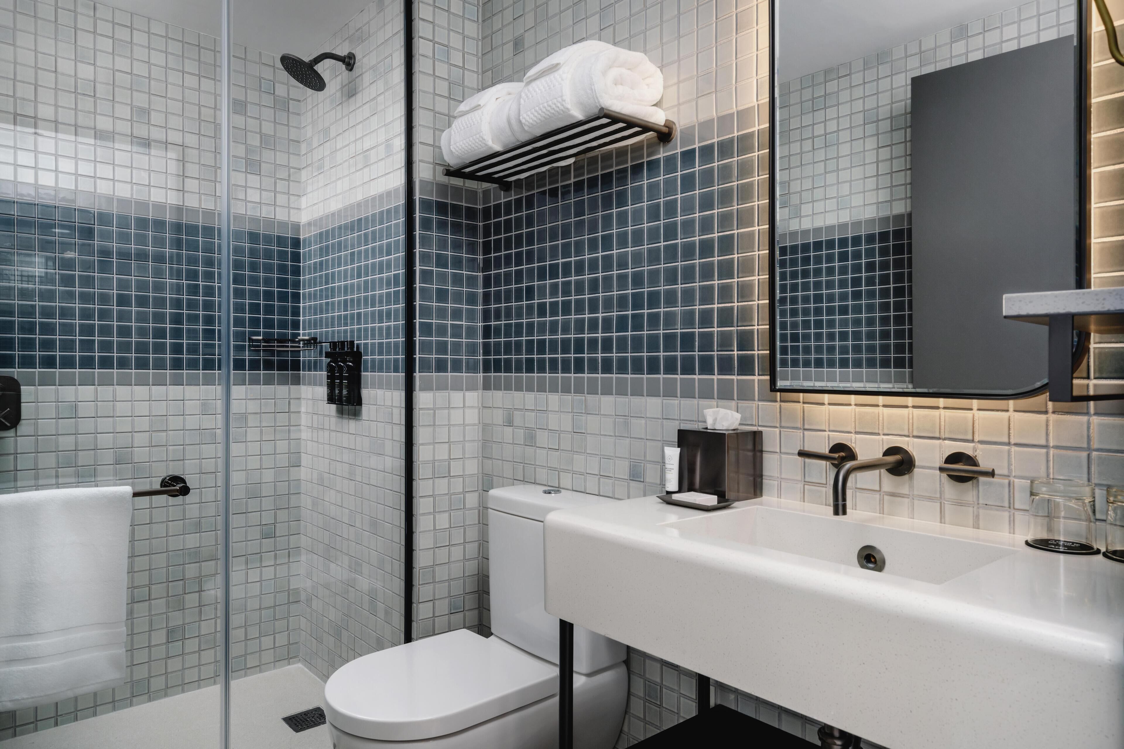 Un baño moderno con cabina de ducha de vidrio, inodoro blanco, lavamanos blanco con espejo y toallero con toallas dobladas.