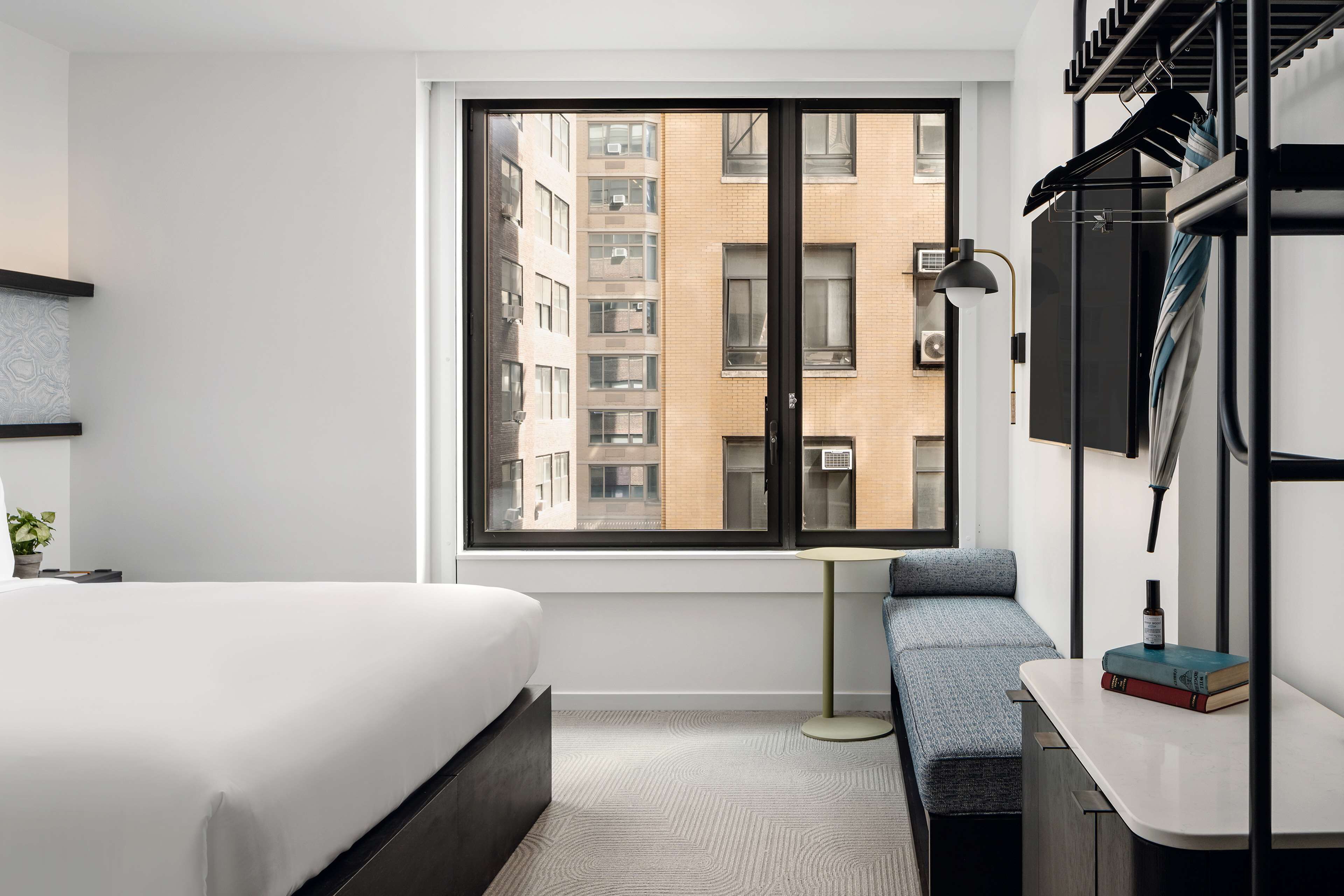 Une chambre d’hôtel moderne dotée d’une grande fenêtre, d’un lit, d’une banquette, d’un bureau et d’un porte-vêtements.