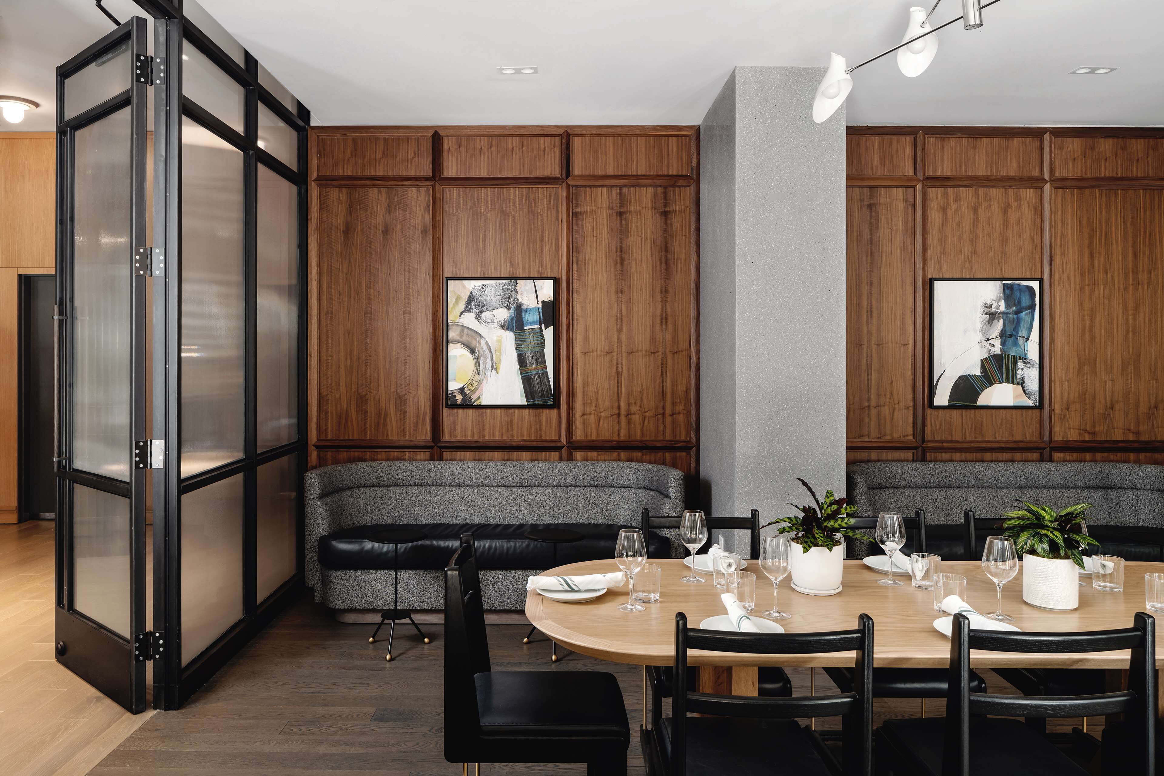 A modern dining area with a round table set for four, surrounded by black chairs. There is a grey cushioned bench against the wall, and two framed abstract paintings are hung above the bench.