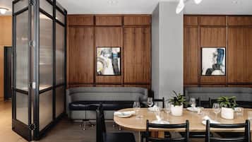 A modern dining area with a round table set for four, surrounded by black chairs. There is a grey cushioned bench against the wall, and two framed abstract paintings are hung above the bench.