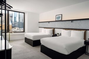 A hotel room with two beds, a large window with a city view, a small table, and a wall-mounted shelf with a picture.