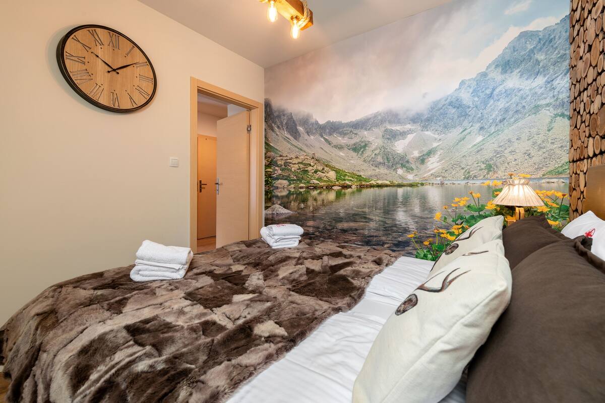 A bedroom with a mountain lake mural, a wooden clock, and a fur throw on the bed.