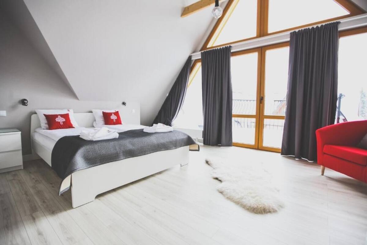 A bedroom with a bed, a red chair, a white nightstand, and a large window with curtains.