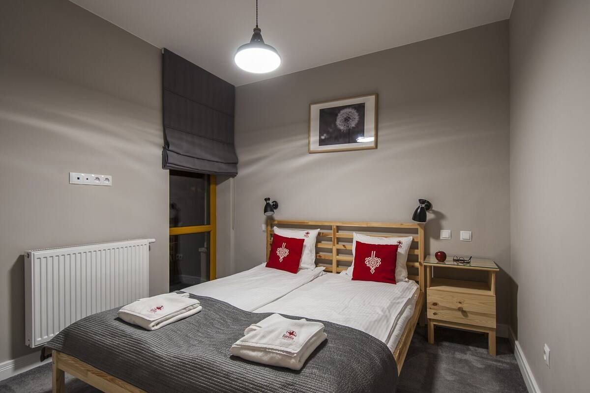 A bedroom with a bed, bedside tables, a radiator, a window with a blind, and a hanging light fixture.