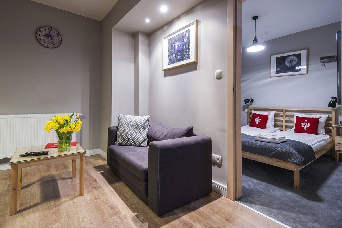 A compact room with a sofa, a small table with a vase of flowers, a bed with red and white pillows, and a clock on the wall.