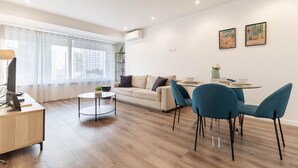 A modern living room with a sofa, dining table, and chairs, a desk with a lamp, and a large window with curtains. - Savory Black Apartment, Sete Rios, Lisbon!New! (Lisboa)