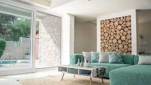 A modern living room with a teal sofa, a glass door leading to a pool, and a wall decorated with stacked logs. - Villa Hvar Beauty with Swimming Pool (Jelsa)