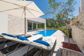 A poolside area with lounge chairs and an umbrella, a stone wall, and a view of greenery.