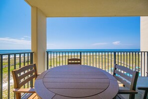 A balcony with a round table and two chairs, overlooking a beach with umbrellas and a clear sky.