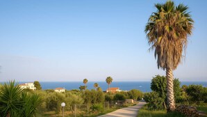 Ein gepflasterter Weg führt zu einer Küstenlandschaft mit Palmen und Häusern im Hintergrund.