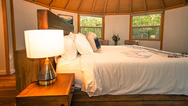 A bedroom with a wooden ceiling, a bed with white bedding, a bedside table with a lamp, and two windows with a view of greenery. - Relax in your own secluded Yurt Cabin nestled into a 200+ acre Property! (Bastrop)