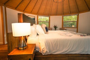 Ein Schlafzimmer mit Holzboden, einem Bett mit weißer Bettwäsche, einem Nachttisch mit Lampe und zwei Fenstern mit Blick auf Grünflächen.