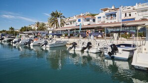 Un port de plaisance où sont amarrés plusieurs bateaux, un ciel dégagé et, au loin, des bâtiments dotés de balcons et de boutiques.