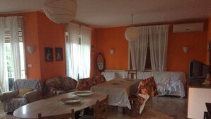 A dining area with a round table, chairs, and a sofa. There are curtains, a television, and a wall-mounted air conditioning unit.