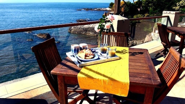 Een houten tafel met een gele tafelkleed, twee stoelen en een bord met eten en drinken op een balkon met uitzicht op zee.
