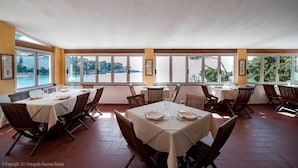 Een eetruimte met tafels voor vier personen, grote ramen met uitzicht op zee en een hoge plafond.