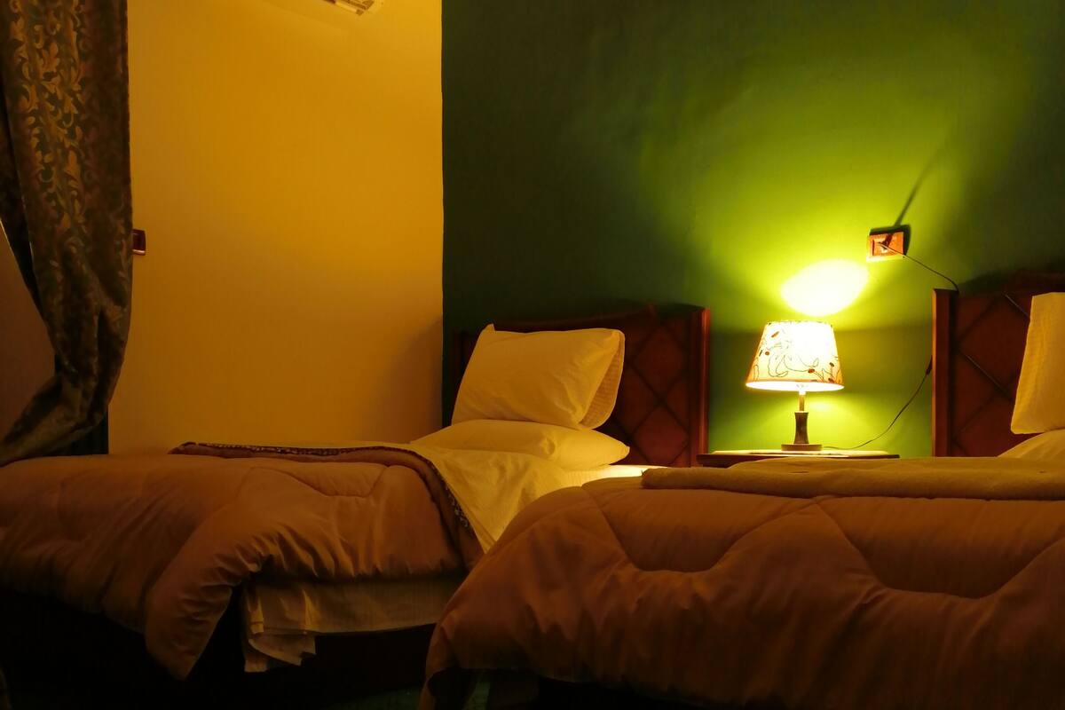 Ein Hotelzimmer mit zwei Betten, einer Klimaanlage, einem Nachttisch mit Lampe und einer Wanduhr.