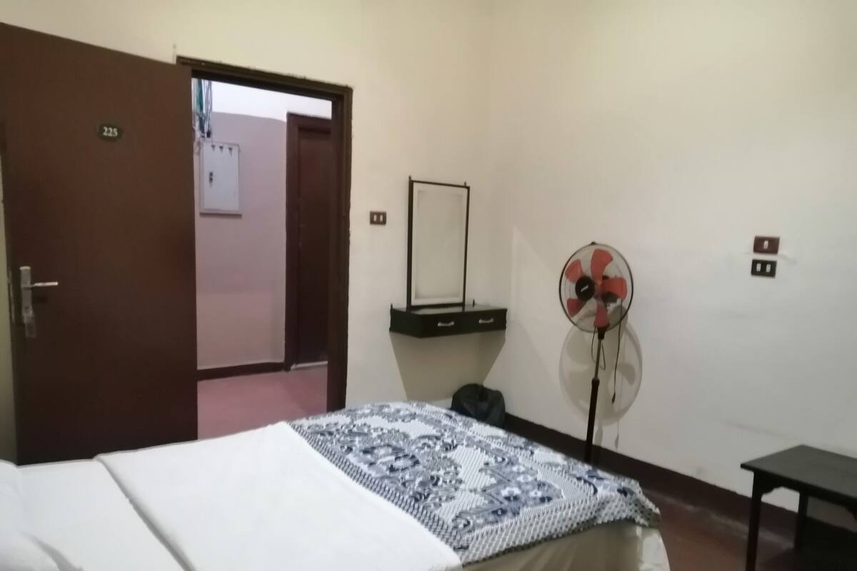 Ein Hotelzimmer mit einem Bett, einem Standventilator, einem Schreibtisch und einer Tür.