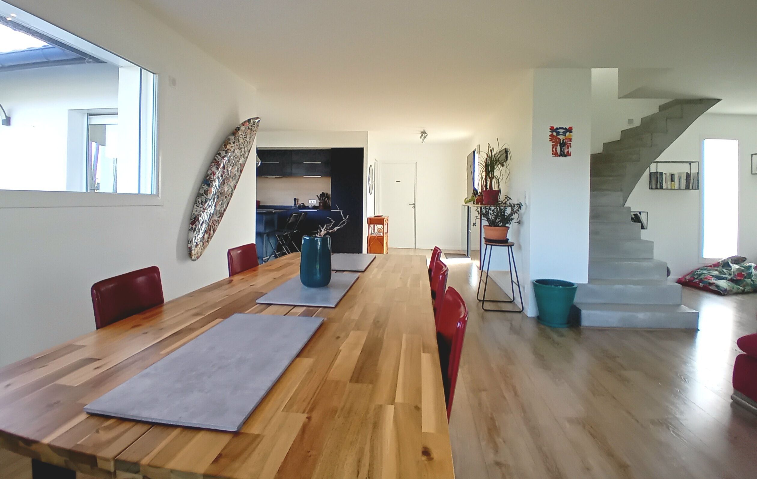 Ruime eethoek met een lange houten tafel en rode stoelen, een keuken achtergrond en een trap naar een ander niveau.