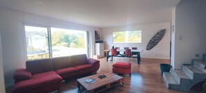 A spacious living room with a red sofa, a wooden coffee table, and a dining area with a table and chairs. Large windows provide natural light and a view of the outdoors.
