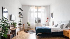 A modern bedroom with a bed, bedside table, and a desk with a chair. There is a window with curtains, a potted plant, and a rug on the floor. - Visitting Apartments - Podwale (Wroclaw)