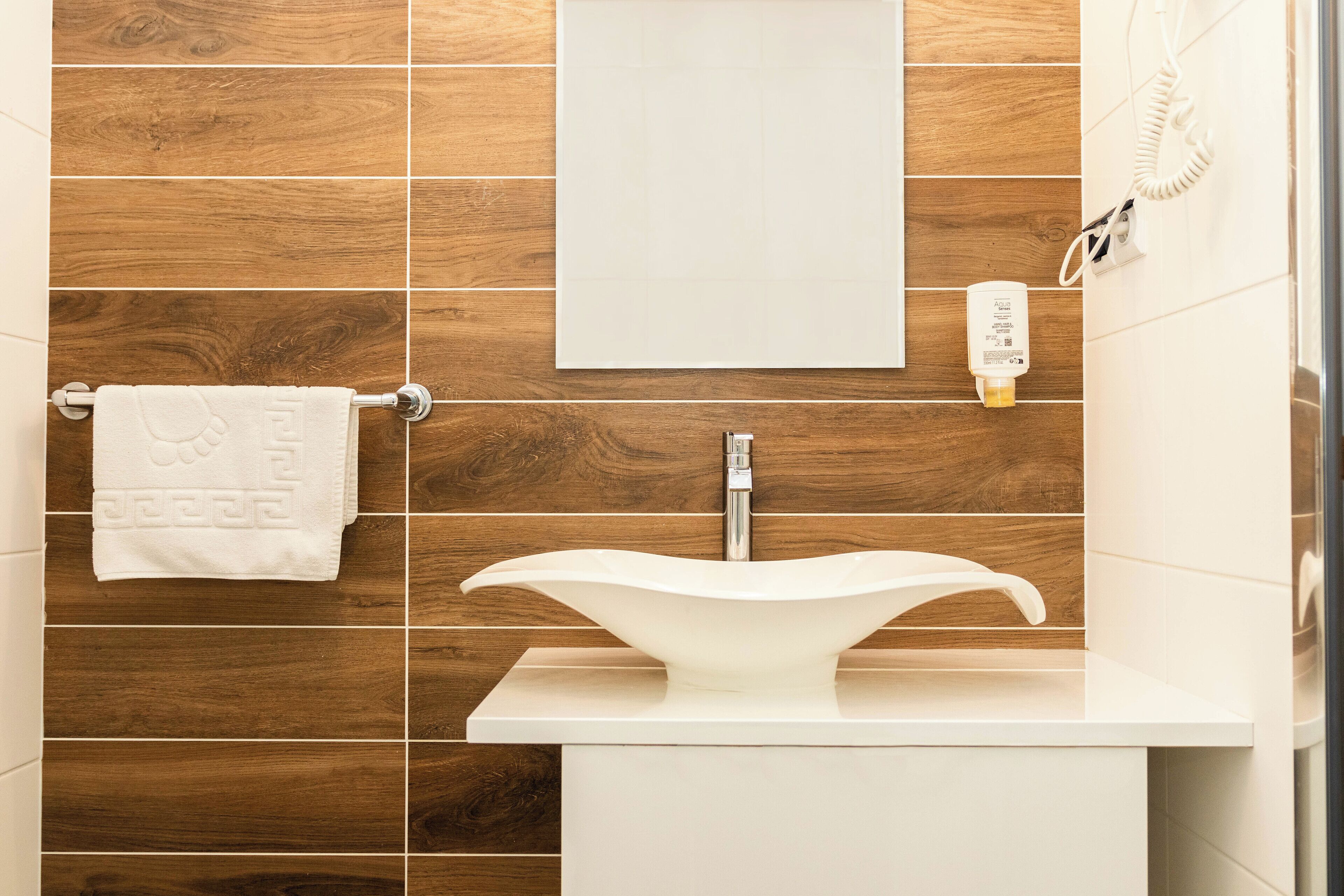A bathroom with a white sink, wooden wall tiles, a towel rack with a towel, a mirror, and a phone charger.
