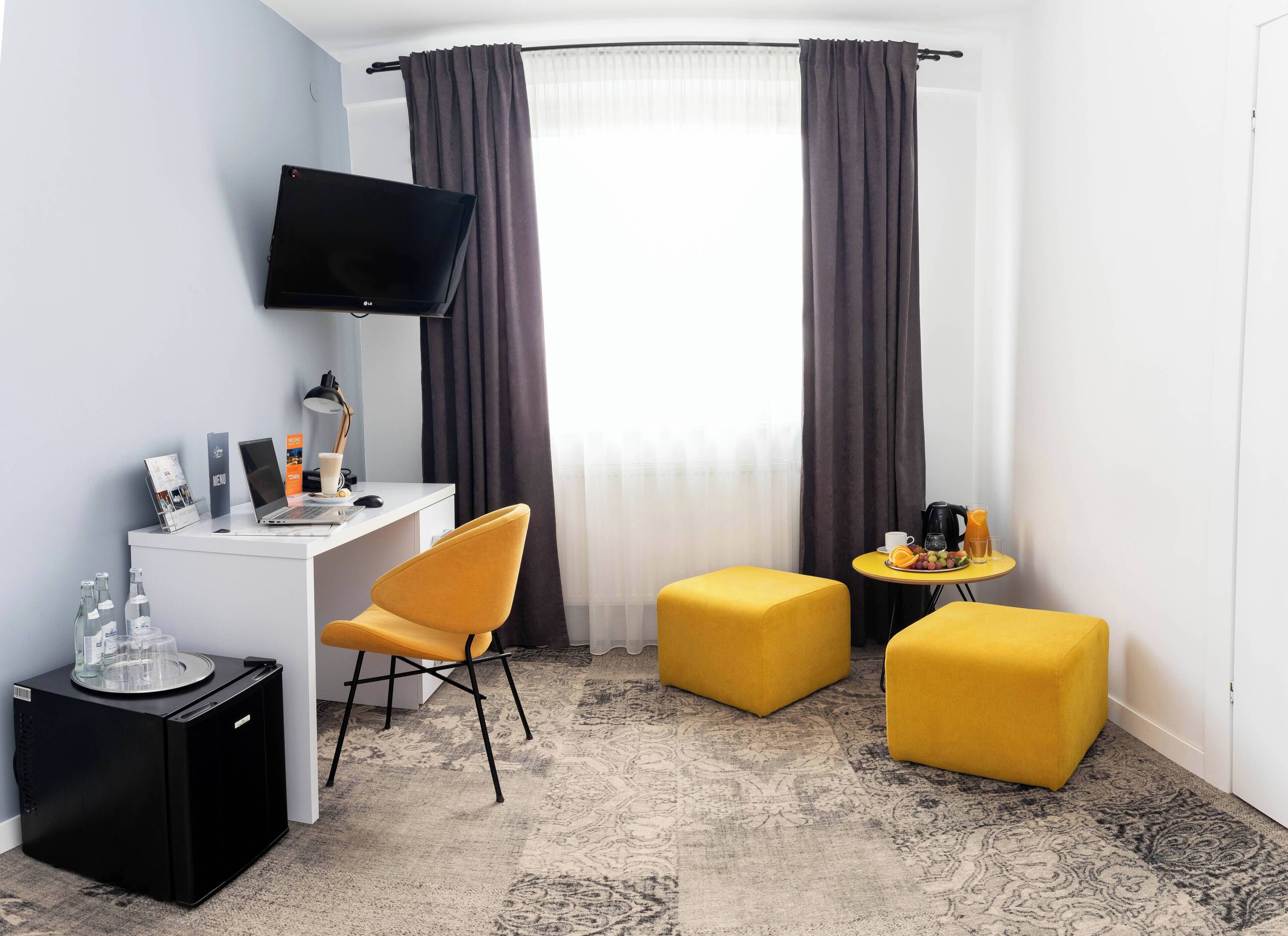 Habitación de hotel compacta con escritorio blanco, televisor de pantalla plana, silla amarilla, dos otomanas amarillas, una pequeña mesa redonda con cafetera y fruta, y un minibár.