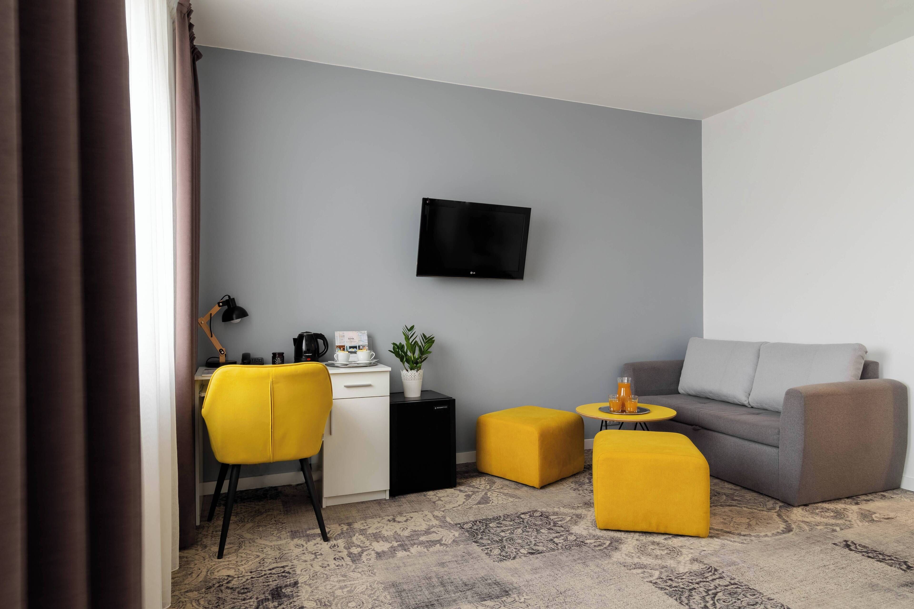 A modern living room with a grey sofa, yellow ottomans, a small coffee table, a desk with a laptop, a plant, and a flat-screen TV mounted on the wall.