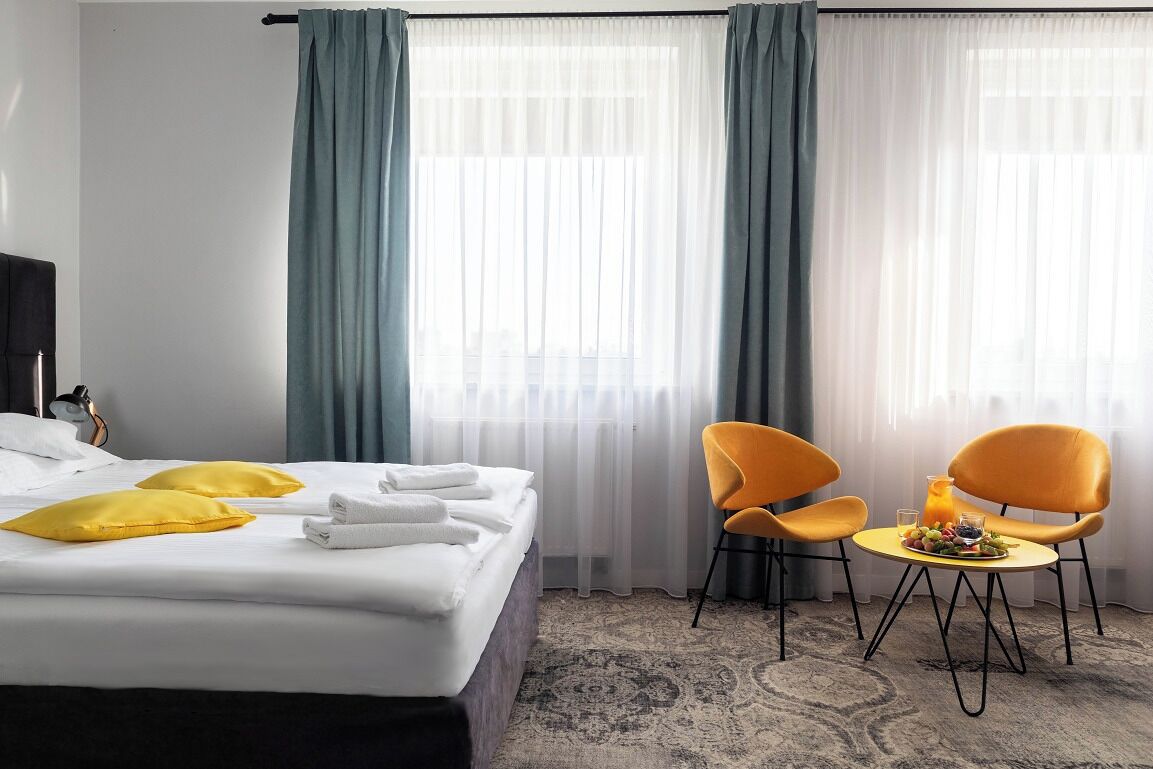 Una habitación de hotel con una cama, dos almohadas amarillas, una mesita de noche con un vaso de zumo de naranja y dos sillones naranjas.