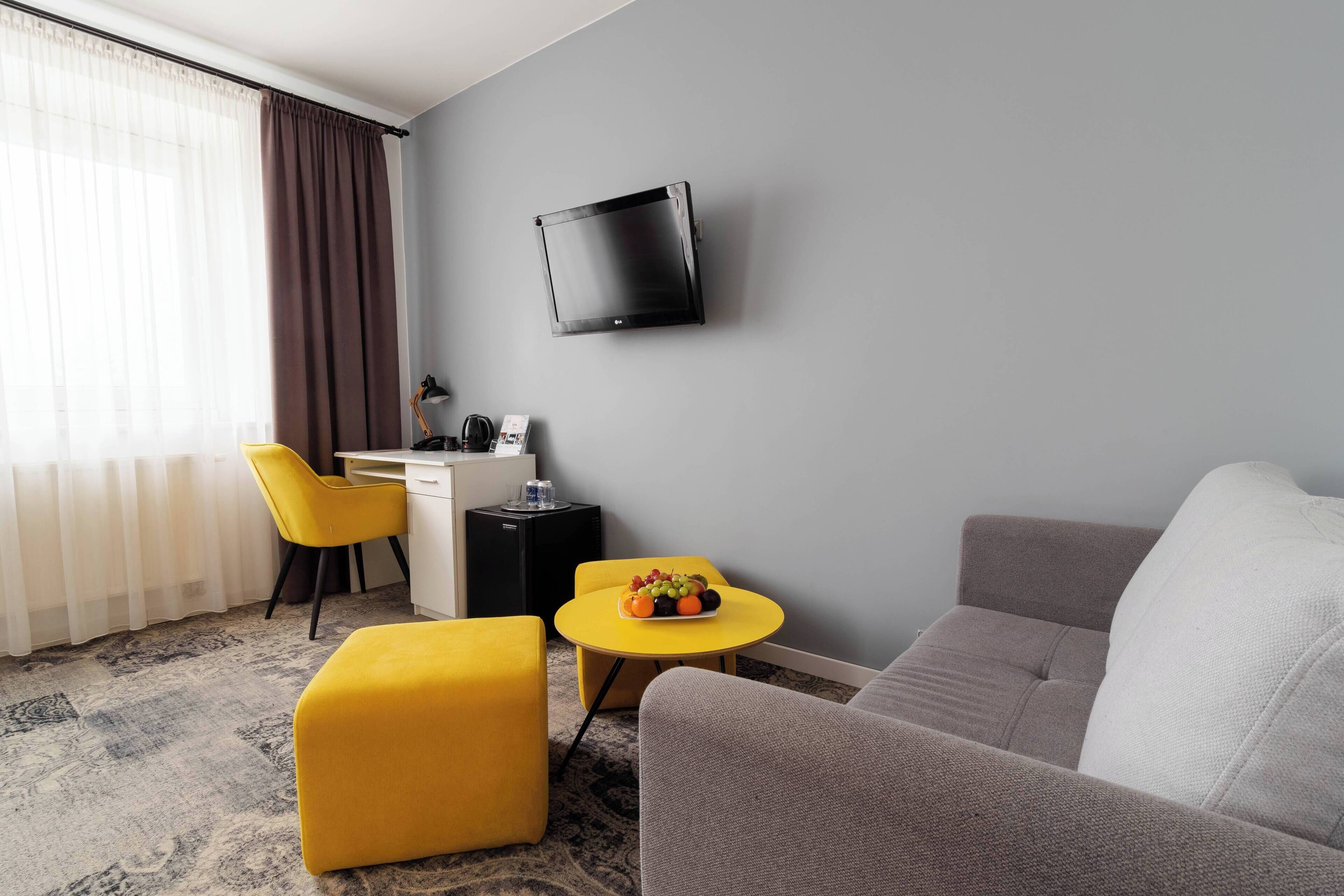 A modern hotel room with a grey sofa, a yellow ottoman, a small round table with fruit, a desk with a chair, and a mounted TV.