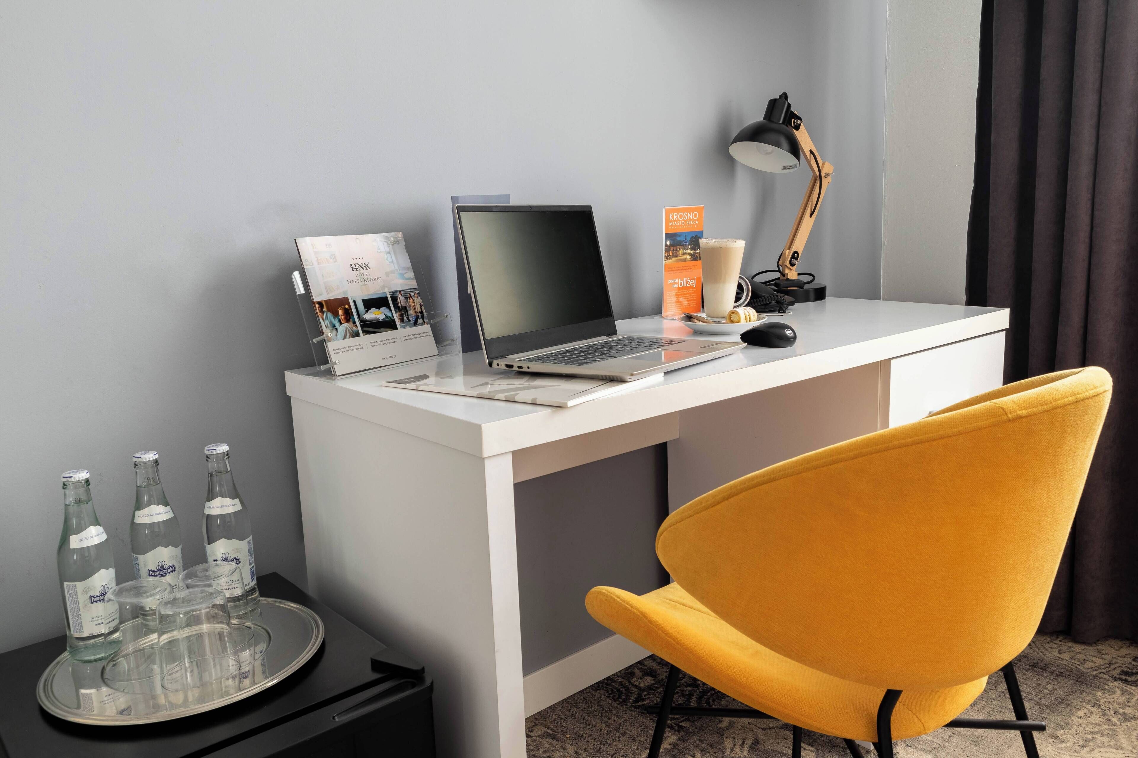 A modern hotel room with a white desk, laptop, and a yellow chair. There are three bottles on a tray, a lamp, and a brochure on the desk.