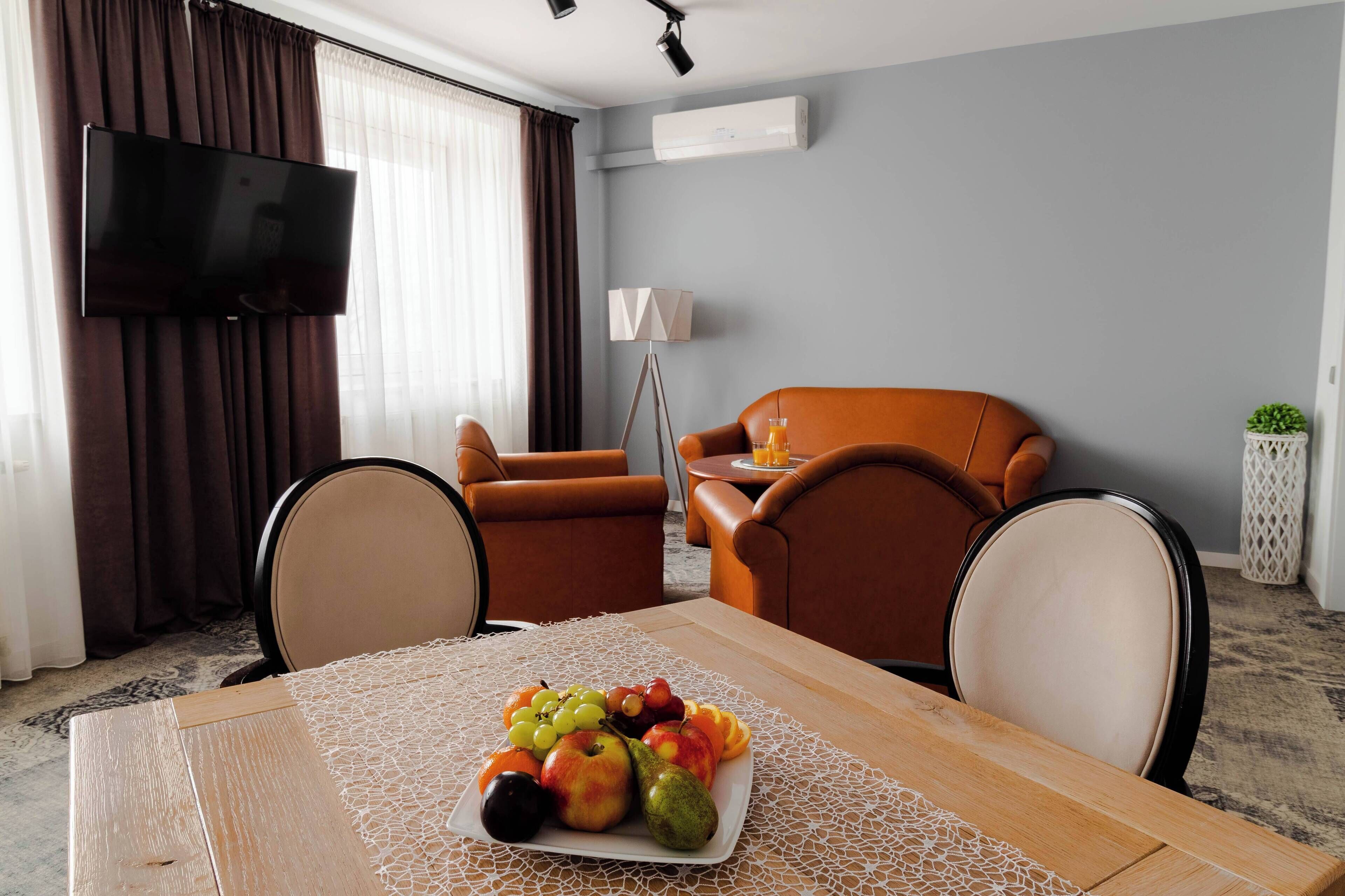 A modern living room with a dining table set with fruit, a flat-screen TV, brown leather sofas, and a lamp.