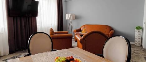 A modern living room with a dining table set with fruit, a flat-screen TV, brown leather sofas, and a lamp.