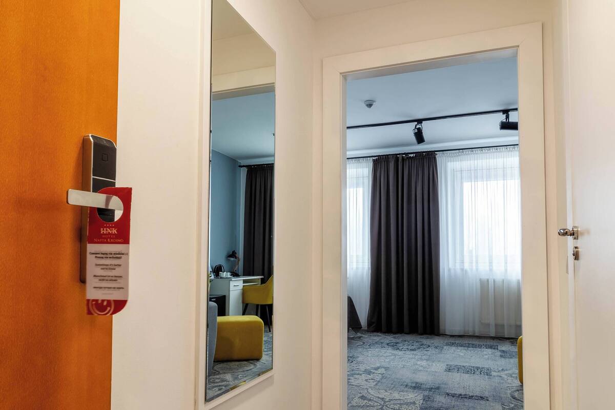 A hotel hallway with a mirror, a door with a lock, a carpeted floor, and a room visible through an open door.