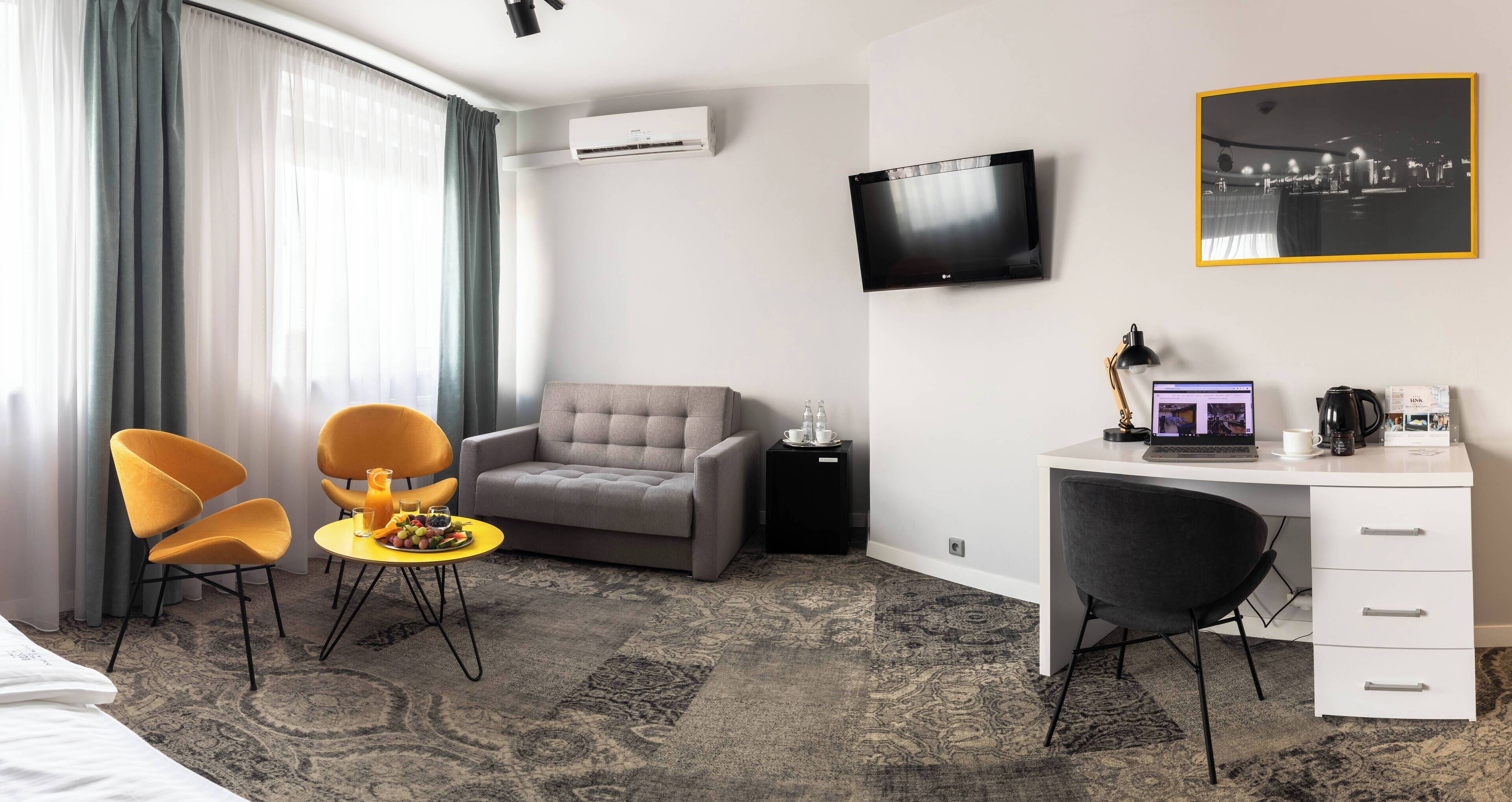 Habitación de hotel con sofá gris, dos sillones amarillos, una mesita redonda con fruta, un escritorio con una computadora portátil, un televisor y un cuadro en la pared.